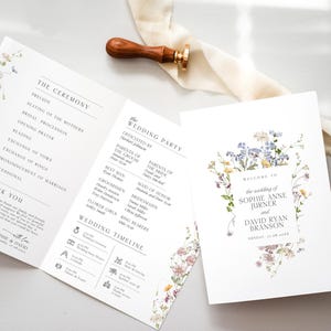 May include: A folded wedding program and a matching invitation card featuring delicate watercolor wildflower illustrations. The text includes the wedding party, ceremony order, and the names Sophie Anne Jurner and David Ryan Branson. A wooden wax seal stamp rests nearby.