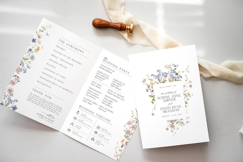 May include: A folded wedding program and a matching invitation card featuring delicate watercolor wildflower illustrations. The text includes the wedding party, ceremony order, and the names Sophie Anne Jurner and David Ryan Branson. A wooden wax seal stamp rests nearby.