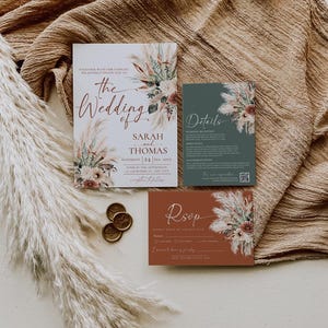May include: A wedding invitation suite featuring a main card, a details card, and an RSVP card. The cards are decorated with dried floral illustrations in muted tones. Two gold wax seals are placed next to the cards on a textured fabric.