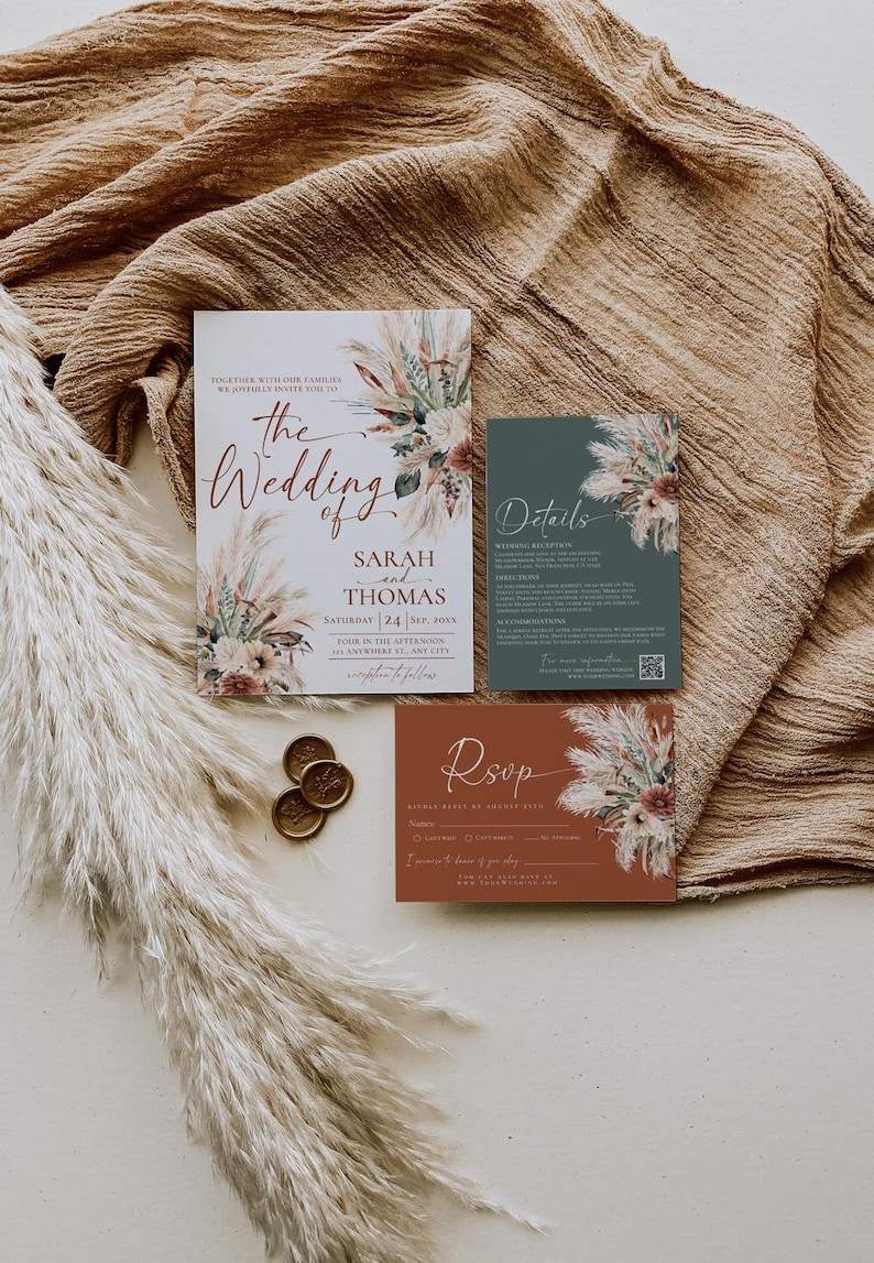 May include: A wedding invitation suite featuring a main card, a details card, and an RSVP card. The cards are decorated with dried floral illustrations in muted tones. Two gold wax seals are placed next to the cards on a textured fabric.