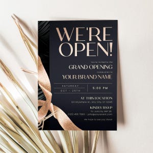 May include: A navy blue invitation with gold text and accents, announcing a grand opening. The text reads "WE'RE OPEN!" and includes event details like the date, time, and location. A gold ribbon design is featured on the invitation.