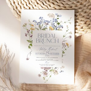 May include: A floral bridal brunch invitation with a watercolor design of blue, yellow, and pink flowers. The text reads "Bridal Brunch" in a light blue font, followed by "Please join us in honor of the bride-to-be Ashley Robert Saturday 25 December 20XX Two o'clock in the afternoon at this location 123 Anywhere St. Any City, ST 12345 Rep On or before November 15th at (123) 45 7890".