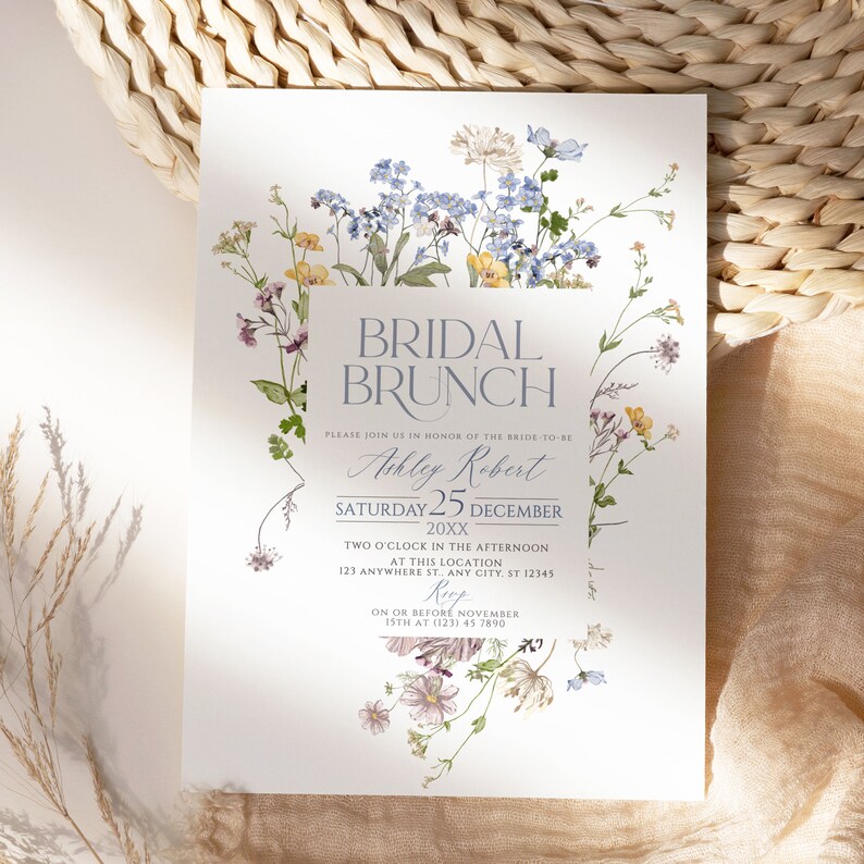 May include: A floral bridal brunch invitation with a watercolor design of blue, yellow, and pink flowers. The text reads "Bridal Brunch" in a light blue font, followed by "Please join us in honor of the bride-to-be Ashley Robert Saturday 25 December 20XX Two o'clock in the afternoon at this location 123 Anywhere St. Any City, ST 12345 Rep On or before November 15th at (123) 45 7890".