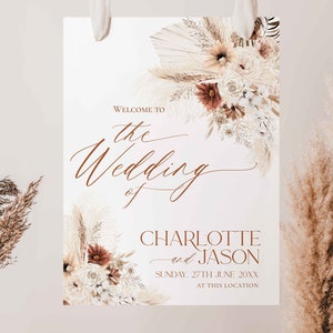 May include: A rectangular wedding welcome sign featuring dried pampas grass and floral arrangements in cream, beige, and rust tones. The text reads: Welcome to the Wedding of Charlotte and Jason, Sunday, 27th June 20xx, at this location.