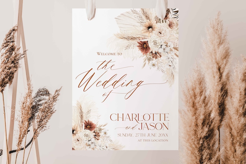 May include: A rectangular wedding welcome sign featuring dried pampas grass and floral arrangements in cream, beige, and rust tones. The text reads: Welcome to the Wedding of Charlotte and Jason, Sunday, 27th June 20xx, at this location.