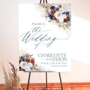 May include: Welcome to the wedding of Charlotte and Jason, a wedding sign with a floral design in shades of blue, white, and rust. The sign is on a wooden easel and the text is in a blue font.