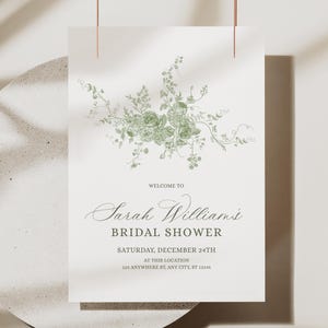May include: A white invitation card featuring a green floral illustration. The text reads "WELCOME TO Sarah Williams BRIDAL SHOWER SATURDAY, DECEMBER 24TH AT THIS LOCATION 123 ANYWHERE ST, ANY CITY, ST 12345". The card is suspended by a thin copper wire.