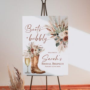 May include: A rectangular sign on a metal easel featuring the text Boots + bubbly and Welcome to Sarah's BRIDAL BRUNCH August 27th 202X. The design includes watercolor illustrations of a brown cowboy boot, a champagne flute, and pampas grass.
