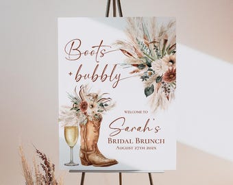 以下が含まれることがあります： 金属製のイーゼルに立てかけられた長方形の看板。Boots + bubbly、Welcome to Sarah's BRIDAL BRUNCH August 27th 202Xという文字が書かれています。茶色のカウボーイブーツ、シャンパングラス、パンパスグラスの水彩画イラストが描かれています。