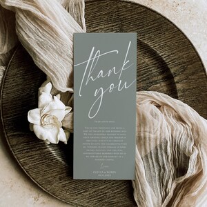 May include: A rectangular sage green thank you card featuring white cursive text that reads Thank you, followed by smaller printed text and the names Olivia & Robin with the date 10.11.20XX at the bottom of the card.