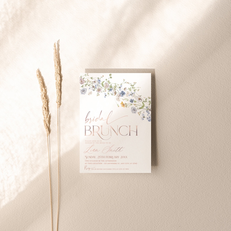 May include: A floral bridal brunch invitation with a watercolour design of blue and yellow flowers. The text reads "bridal brunch" in a rose gold font, followed by "Please join us in honour of the bride-to-be, Liem Smith. Sunday, 25th February 20XX. Two o'clock in the afternoon. At The Location, 121 Anywhere St, Any City, ST 12345."
