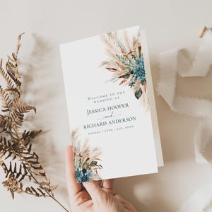 May include: A folded wedding invitation with floral and pampas grass accents. The invitation reads "Welcome to the Wedding of Jessica Hooper and Richard Anderson" with the date below. The design features a watercolor style with muted colors.