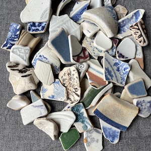 May include: A pile of broken ceramic fragments, showcasing a variety of patterns and colours. The pieces feature blue, white, green, and brown hues, with floral and geometric designs. Some shards have curved edges, indicating they are from plates or bowls.
