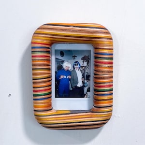 Może przedstawiać: Prostokątna ramka na zdjęcia wykonana z warstwowego drewna, z kolorowym wzorem w paski. Ramka otacza zdjęcie w stylu polaroid dwóch osób w kostiumach. Ramka jest zamontowana na białej ścianie.