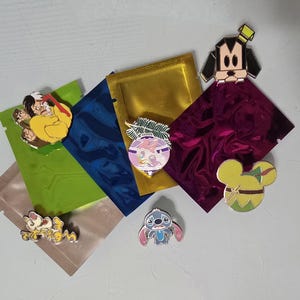 May include: Collection of enamel pins featuring cartoon characters, including Snow White, Goofy, Stitch, and Mickey Mouse. The pins are displayed on colorful metallic pouches. The pins have various colors, including yellow, blue, pink, and green.