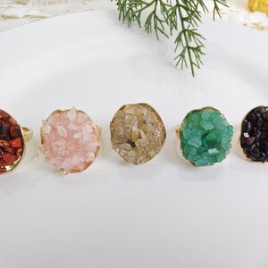 Op de afbeelding: Vijf edelsteenringen met goudkleurige banden worden gepresenteerd op een wit oppervlak. De edelstenen zijn rood, roze, bruin, groen en donkerpaars. Elke ring heeft een cluster van kleine, onregelmatig gevormde stenen.