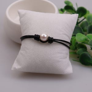 May include: A black leather bracelet with a single white pearl accent. The bracelet is displayed on a white pillow.