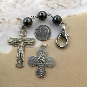 Puede incluir: Un rosario de plata con cuentas negras y un crucifijo de plata. El crucifijo presenta una representaci&oacute;n de Jesucristo. El rosario tambi&eacute;n incluye un dije de plata con las palabras "Soy cat&oacute;lico, por favor llama a un sacerdote".