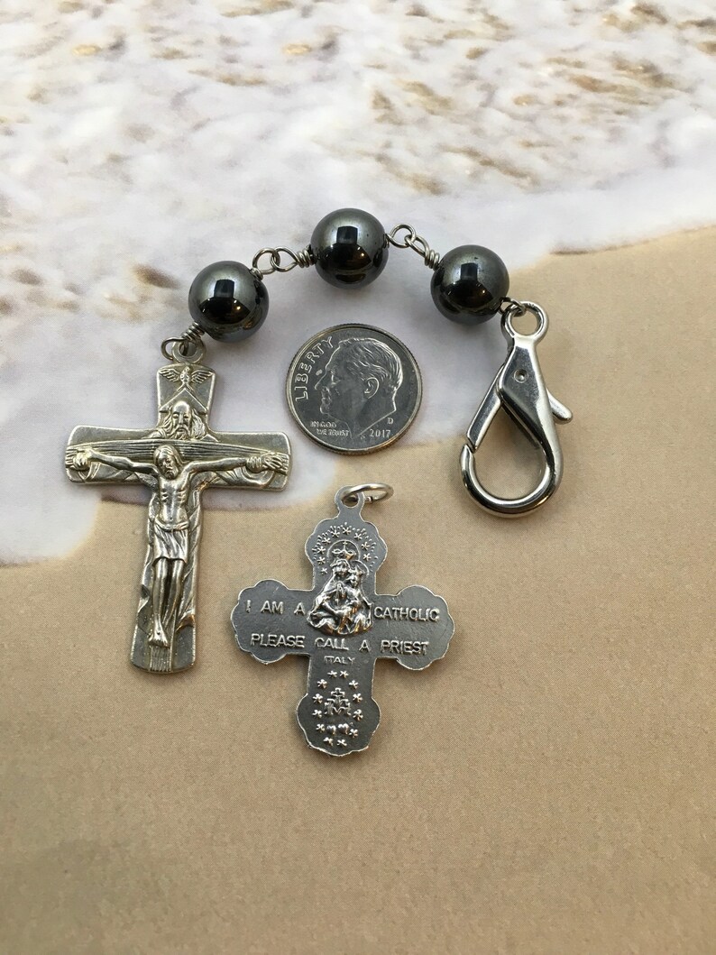 Puede incluir: Un rosario de plata con cuentas negras y un crucifijo de plata. El crucifijo presenta una representaci&oacute;n de Jesucristo. El rosario tambi&eacute;n incluye un dije de plata con las palabras "Soy cat&oacute;lico, por favor llama a un sacerdote".