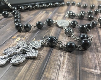 May include: A black hematite rosary with a silver crucifix and a silver medal. The rosary beads are round and have a shiny finish. The crucifix is ornate and features a figure of Jesus Christ. The medal is circular and has a religious image.