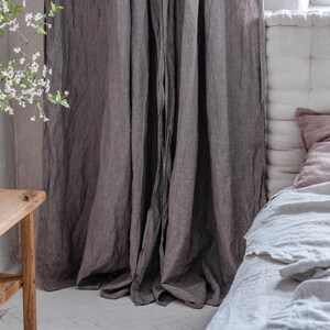 May include: Gray linen curtains hanging in a bedroom with a white bed and a wooden side table. The curtains are gathered at the top and have a natural, relaxed look.
