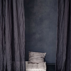 Op de afbeelding: Een wit, gestoffeerd vloer kussen met een grijs kussen voor een donkergrijze muur. Het kussen is in een kamer met twee donkergrijze gordijnen aan beide kanten.