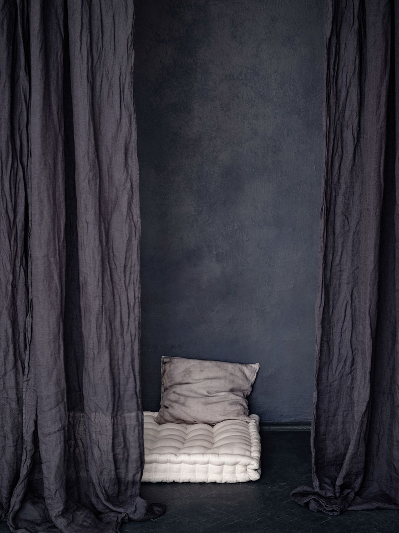 Op de afbeelding: Een wit, gestoffeerd vloer kussen met een grijs kussen voor een donkergrijze muur. Het kussen is in een kamer met twee donkergrijze gordijnen aan beide kanten.