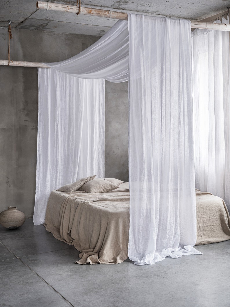 May include: A bed with a white linen canopy and a beige linen duvet. The bed is in a room with a concrete floor and a white wall.