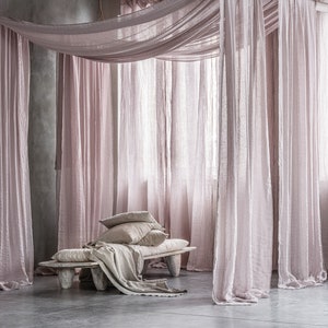 May include: A light pink linen canopy draped over a wooden bench with white linen pillows. The bench is in a room with a concrete floor and walls.