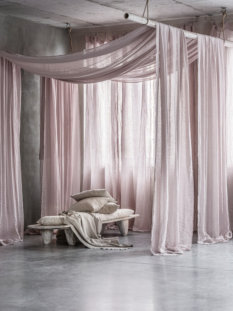 May include: A light pink linen canopy draped over a wooden bench with white linen pillows. The bench is in a room with a concrete floor and walls.