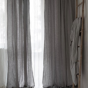 May include: Two sets of gray linen curtains hang in a window, with a white sheer curtain behind them. A wooden ladder leans against a white wall with a gray linen throw draped over it.