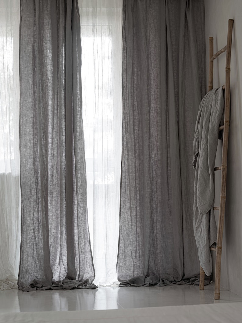 May include: Two sets of gray linen curtains hang in a window, with a white sheer curtain behind them. A wooden ladder leans against a white wall with a gray linen throw draped over it.
