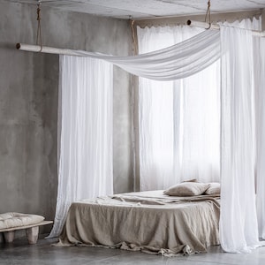 May include: A bed with a white linen canopy and a beige linen duvet cover. The bed is in a room with a concrete wall and a wooden bench.