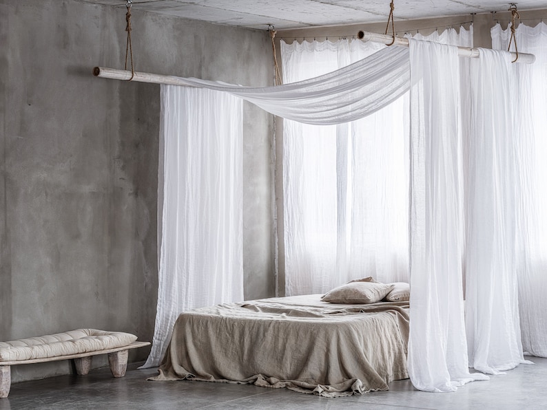 May include: A bed with a white linen canopy and a beige linen duvet cover. The bed is in a room with a concrete wall and a wooden bench.