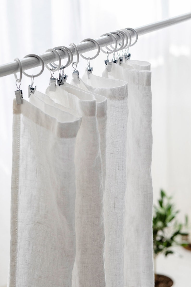 May include: White linen curtains hanging on a white curtain rod with silver clips.