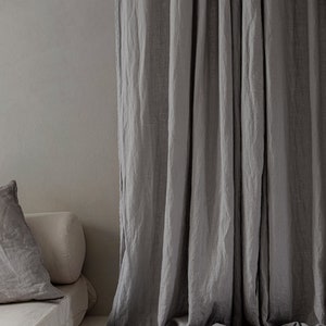 K&ouml;nnte beinhalten: Graue Leinenvorh&auml;nge, die in einem Raum mit einer Couch und Kissen h&auml;ngen. Die Vorh&auml;nge sind aus einem weichen, nat&uuml;rlichen Stoff gefertigt und haben einen entspannten, l&auml;ssigen Look.