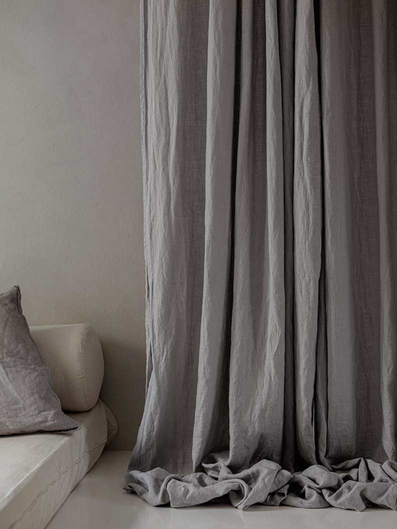 K&ouml;nnte beinhalten: Graue Leinenvorh&auml;nge, die in einem Raum mit einer Couch und Kissen h&auml;ngen. Die Vorh&auml;nge sind aus einem weichen, nat&uuml;rlichen Stoff gefertigt und haben einen entspannten, l&auml;ssigen Look.