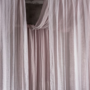 May include: A pair of sheer, light pink curtains hang from a wooden rod. The curtains are gathered at the top and drape down in soft folds.