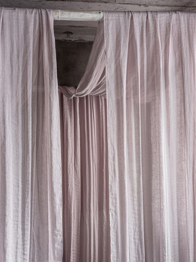 May include: A pair of sheer, light pink curtains hang from a wooden rod. The curtains are gathered at the top and drape down in soft folds.