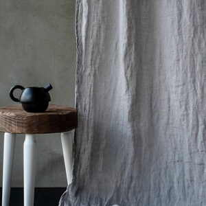 Op de afbeelding: Een grijs linnen gordijn hangt voor een betonnen muur. Een zwarte theepot staat op een houten kruk met witte poten.