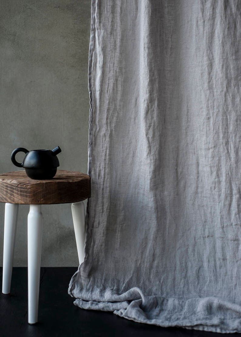 Op de afbeelding: Een grijs linnen gordijn hangt voor een betonnen muur. Een zwarte theepot staat op een houten kruk met witte poten.