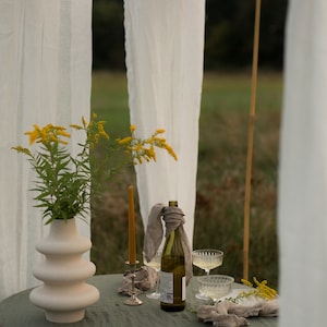 May include: A green tablecloth with a white vase filled with yellow flowers, a bottle of wine, two glasses, and a candle holder. The table is set up outdoors under a white canopy.