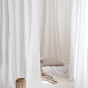 May include: A white linen canopy bed with a brown pillow and white sheets. A wicker lantern sits on the white floor. Four white candles are on a wooden base.