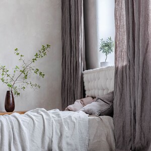 K&ouml;nnte beinhalten: Ein wei&szlig;es Leinenbett mit einem gesteppten Kopfteil und einer grauen Leinen-&Uuml;berwurfdecke. Das Bett ist mit einem hellrosa Kissen und einem grauen Kissen bezogen. Das Bett steht in einem Raum mit grauen Leinenvorh&auml;ngen und einer kleinen Topfpflanze.
