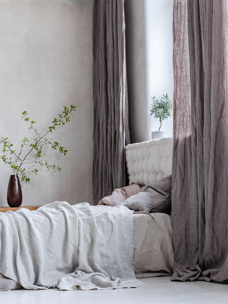 K&ouml;nnte beinhalten: Ein wei&szlig;es Leinenbett mit einem gesteppten Kopfteil und einer grauen Leinen-&Uuml;berwurfdecke. Das Bett ist mit einem hellrosa Kissen und einem grauen Kissen bezogen. Das Bett steht in einem Raum mit grauen Leinenvorh&auml;ngen und einer kleinen Topfpflanze.