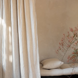 K&ouml;nnte beinhalten: Ein hellbeiger Leinenvorhang h&auml;ngt vor einer hellbraunen Wand. Im Hintergrund befindet sich ein Futon mit einer hellbeigen Leinenmatratze und zwei Kissen. Ein Zweig mit roten Beeren befindet sich in der Ecke des Bildes.