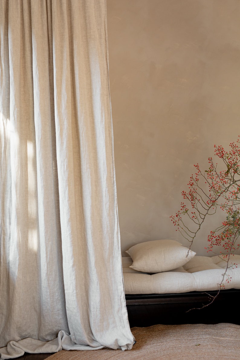 K&ouml;nnte beinhalten: Ein hellbeiger Leinenvorhang h&auml;ngt vor einer hellbraunen Wand. Im Hintergrund befindet sich ein Futon mit einer hellbeigen Leinenmatratze und zwei Kissen. Ein Zweig mit roten Beeren befindet sich in der Ecke des Bildes.