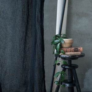 Op de afbeelding: Een zwart, doorzichtig gordijn hangt voor een grijze muur. Een kleine plant in een pot met groene bladeren staat op een zwarte verstelbare kruk. De kruk is bedekt met een stapel boeken.