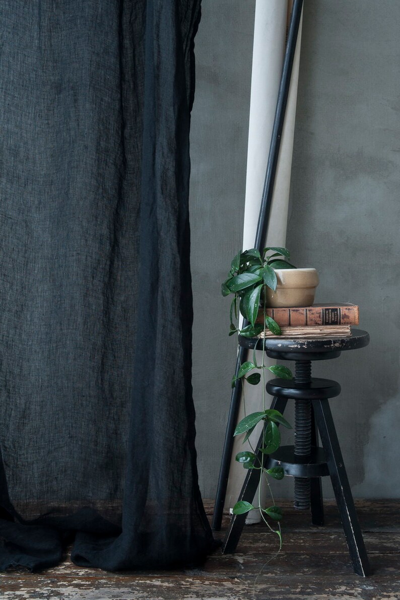 Op de afbeelding: Een zwart, doorzichtig gordijn hangt voor een grijze muur. Een kleine plant in een pot met groene bladeren staat op een zwarte verstelbare kruk. De kruk is bedekt met een stapel boeken.