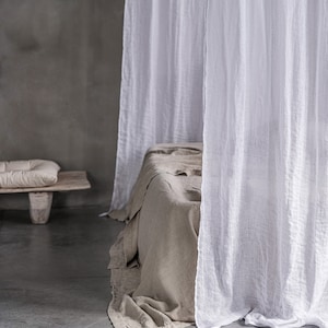 May include: A white linen canopy draped over a bed with a light brown linen duvet. The bed is in a room with a concrete floor and a grey wall.